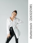 Small photo of fashionable woman in black tight pants and white oversize shirt posing with hand on hip isolated on grey