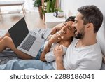 laughing man hugging african american girlfriend while watching film on laptop on bed at home