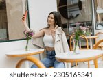 Small photo of happy woman with long hair sitting on chair near bistro table with flowers in vase and taking selfie, shoving v sign, posing in trendy clothes in cafe on terrace outdoors in Istanbul