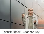 smiling young woman with bangs holding paper cup with coffee to go and standing in trendy outfit and wireless earphones while using smartphone near grey modern building on urban street, look at camera