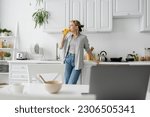 Small photo of tattooed woman in eyeglasses drinking orange juice and standing near kitchen worktop next to desk with devices, bowl with cornflakes and cup of coffee with saucer at home, freelance lifestyle