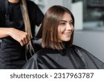 Small photo of client satisfaction, cheerful woman with short brunette hair sitting in hairdressing cape in beauty salon, getting haircut by professional hairdresser, beauty salon