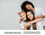Small photo of Gleeful and teenage girls in white t-shirts hugging each other and looking at camera while standing isolated on grey, energetic teenage friends spending time, copy space, friendship and companionship