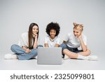 Small photo of Teen african american girl using laptop near girlfriends in white t-shirts and jeans while sitting together on grey background, teenagers bonding over common interest, friendship and companionship
