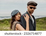 Small photo of Portrait of trendy brunette woman in newsboy cap and vest hugging boyfriend in sunglasses and jacket while looking at camera with landscape at background, trendy twosome in rustic setting