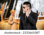 Small photo of bodyguard concept, handsome man in formal wear and tie touching earpiece in lobby of hotel, security, communication, private safety, protecting female client on blurred background
