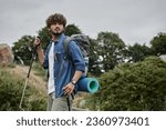 curly indian backpacker holding trekking poles on path in woods, hiker having adventure concept