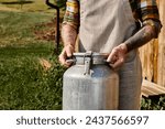 Small photo of cropped view of adult farmer with tattoos on arms holding metal milk churn while in village