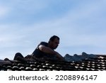Man reparing roof tiles with cement on an old building roof in the village on a sunny day. Silhouette of a man on a roof while working