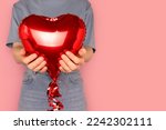 Unrecognizable woman hold in hands red inflatable foil balloon in a heart shape in front of pink background.