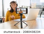Woman radio host in headphones streaming live video podcast using microphone and laptop in studio. Female youtuber looking at camera, shooting vlog. Selective focus on smartphone camera. Close-up