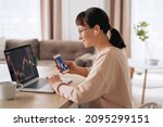 Smiling business woman trader analyst looking at laptop monitor, holding smartphone, wearing earphones. Investor broker analyzing indexes, trading online investment data on stock market graph at home