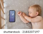 Small photo of Upper view of baby napping in his bed next to phone playing lullaby in app for baby sleep with child tracker function and parents remote control settings. Maternity and technology. Healthy sleep