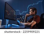 Side view of concentrated crypto trader sitting in front of computers, making professional analysis of candlestick chart, creating strategy, holding pen and notepad, looking at monitor, touching chin