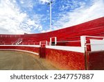 Small photo of Red, wooden bleachers circumvent a brown sandy arena with blue sky and white clouds.