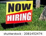 Small photo of Sign text closeup for help wanted now hiring in English with red and yellow colors as signpost on grass for store shop business during corona virus covid 19 pandemic