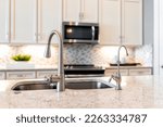 New modern faucet and kitchen sink closeup with island and granite countertops with bokeh background of microwave and oven stove with bright white cabints and nobody