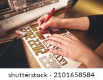 woman with red painted nails removes excess material with weeding tool to make 2022 tiger design stickers from gold vinyl foil. workplace with cutting machine, tools, tablet computer. selective focus