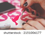 Small photo of women hands with painted nails hold freshly cut peace dove decal that sticks to tip of curved weeding tool. pink adhesive film. wooden worktop. adjustable plotting blade in foreground. selective focus