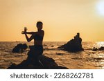 Silhouette of young woman sitting on rocks by the sea in yoga pose against the backdrop of sunset on summer.