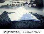 Inspirational motivational quote - Your next chapter is going to be amazing. With open book page and white paper pages flower shape decoration on a sea rock outdoor. White bokeh light background.