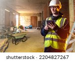 Small photo of Repairman is holding tablet. Man in unfinished building. Construction company employee. Guy in construction vest and helmet. Blurred unfinished building behind builder. Work in construction business