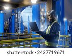 Small photo of Man in industrial factory. Engineer at plant site. Guy is worker at chemical factory. Engineer with laptop in yellow vest. Industrial pressure tanks behind man. Industrialist in modern factory