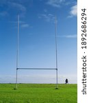Small photo of Tall goal post for Irish National sport rugby, hurling, gaelic football and camogie on a green training pitch, blue cloudy sky. Couple walking in the background