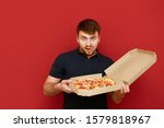 A funny man with a beard holds a pizza box in delivery and looks into the camera with a hungry lustful face. Portrait of hungry man with pizza box on red background. Fast junk food concept.