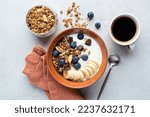 Yogurt with granola, banana, blueberries and dark chocolate chunks in a bowl served with cup of black coffee. Top view healthy breakfast meal