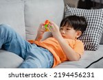 Small photo of A cute Asian boy is lying on the sofa, in his hands he works a small square. Learn to play with the same colors. with intention Emphasize the placement of the same colors in the same row.