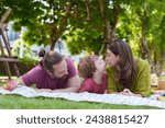 Small photo of Parents leaning in to kiss young boy on picnic blanket, amidst a spread of snacks, portraying family affection and shared joy. Mother and father bestow peck on cheek of boy amidst outdoor feast