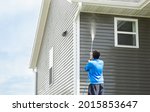 Small photo of Man in blue jacket cleans dusk and dirt from exterior siding and under roof with a high-pressure nozzle spray with water soap cleaner. Wash a house during the day. Home maintenance service concept.