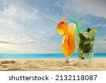 Orange and green cocktails on the beach. Alcoholic drink with ice, orange, mint and lime on the sand. Sunset, sea and sky in the background. Copy space and free space for text near the glass.