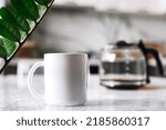 Mock-up of a white mug with coffee or tea is on the table with a glass teapot.