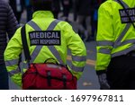 Medical first responders walk along a road wearing black wool stocking caps, and yellow reflective coats with the medical first responder in grey letters and across.The EMT is carrying a first aid kit