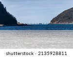 Small photo of A wide view of frozen ice or slob in a cove. This ice has patches of snow with a glass surface. There are mountains or hills in the distance that are covered in green evergreen trees with blue sky.