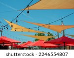 Small photo of Multiple triangle shaped yellow nylon sunshades and awnings hanging over a patio deck. There are red colored canvas umbrellas hung with strings of clear patio light against a bright blue sunny sky.