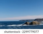 The deep blue Atlantic Ocean with waves crashing on a rocky island. The sea's coastline has rugged high cliffs with hills covered in hills. The sky is blue with a pink hue in the low lying clouds. 