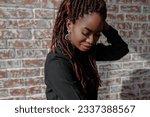 Small photo of A portrait of pretty african woman with flawless skin, dreadlocks hairdo and stylish makeup, wearing black shirt and eloquent earrings, looking down against a brick wall with copy space.