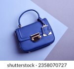 Small photo of Close-up photo of a top-handle royal blue leather women's bag with metal hardware including a clasp, tag, and protective feet, set against a geometric blue-purple background.