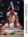 Small photo of Cheerful and happy hellion schoolgirl sitting at the desk with books, school supplies, with a red apple on the top of her head.