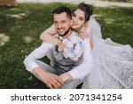 A beautiful, sweet bride in a dress hugs behind the back of a stylish, bearded, smiling groom in a gray vest and white shirt sitting on the green grass. Wedding portrait, photo of newlyweds in love.