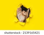 Small photo of Close-up of a woman's ear and hand through a torn hole in the paper. Bright yellow background, copy space. The concept of eavesdropping, espionage, gossip and tabloids.