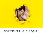 Small photo of Close-up of a left woman's ear and hand through a torn hole in the paper. Bright yellow background, copy space. The concept of eavesdropping, espionage, gossip and tabloids.