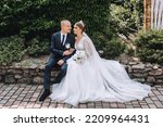 A stylish, young groom and a beautiful bride in a long white dress with a bouquet in their hands, a diadem on their heads, sit gently hugging in a park in nature. Wedding photography of the newlyweds.