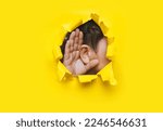 Small photo of Close-up of a man's ear and right hand through a torn hole in the paper. Yellow background, copy space. The concept of eavesdropping, espionage, gossip and tabloids.