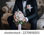 A stylish groom in a blue suit with a boutonniere with a beautiful bouquet in his hands sits on a sofa in a room indoors. Wedding photography, portrait.