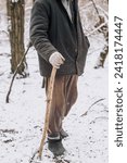 Small photo of An old, elderly, homeless beggar man, a pensioner in dirty clothes, walks through the forest in nature with a wooden stick in search of food, shelter in the snowy winter in the cold. Poverty concept.