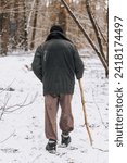 Small photo of An old, elderly forester, a homeless man in dirty clothes walks through the forest with a wooden stick in search of food, shelter in the cold snowy winter.